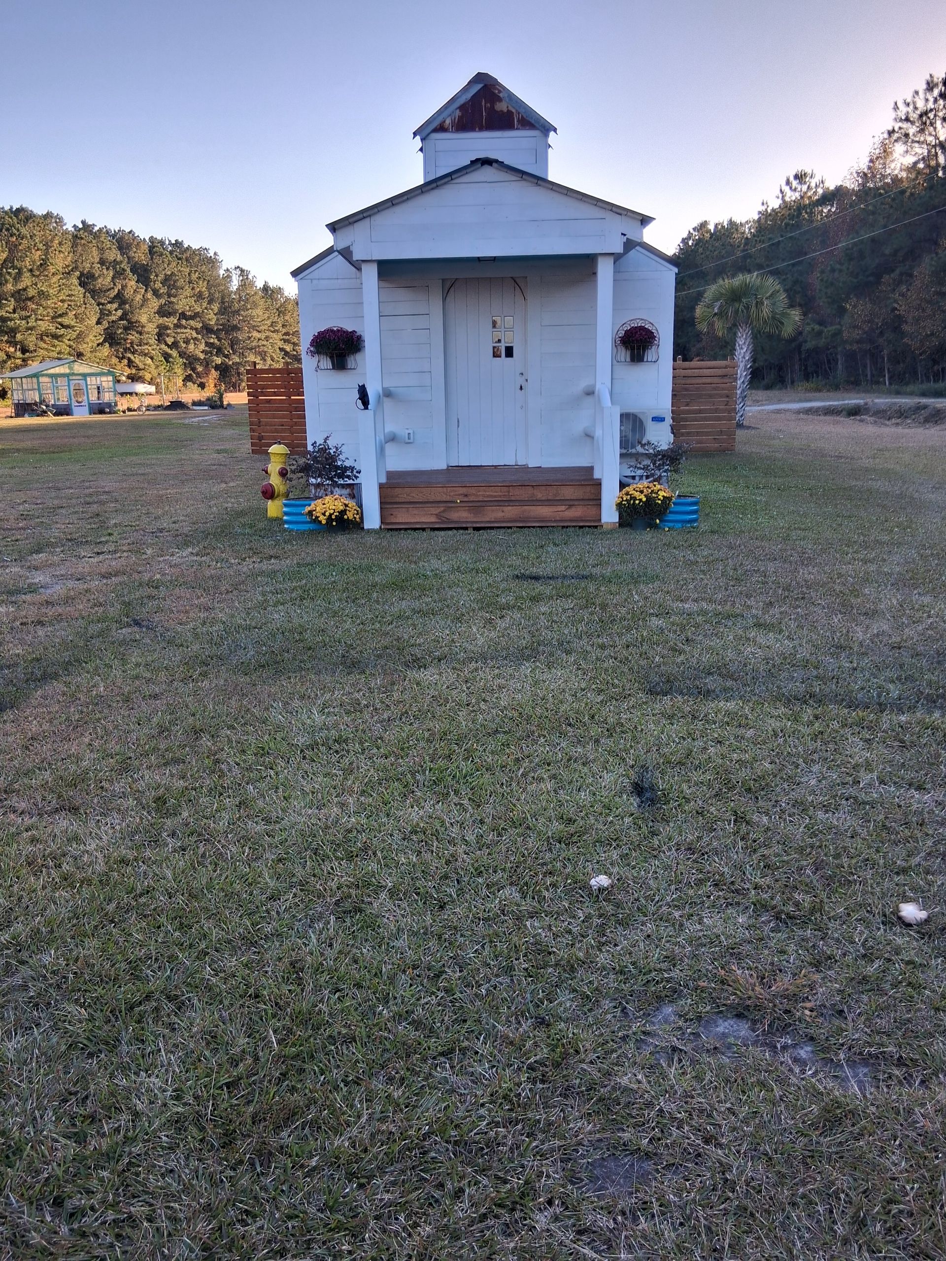 A small, white, chapel-style shed stands in a grassy field with floral accents and wooden steps at the entrance. A small, white, chapel-style shed stands in a grassy field with floral accents and wooden steps at the entrance.