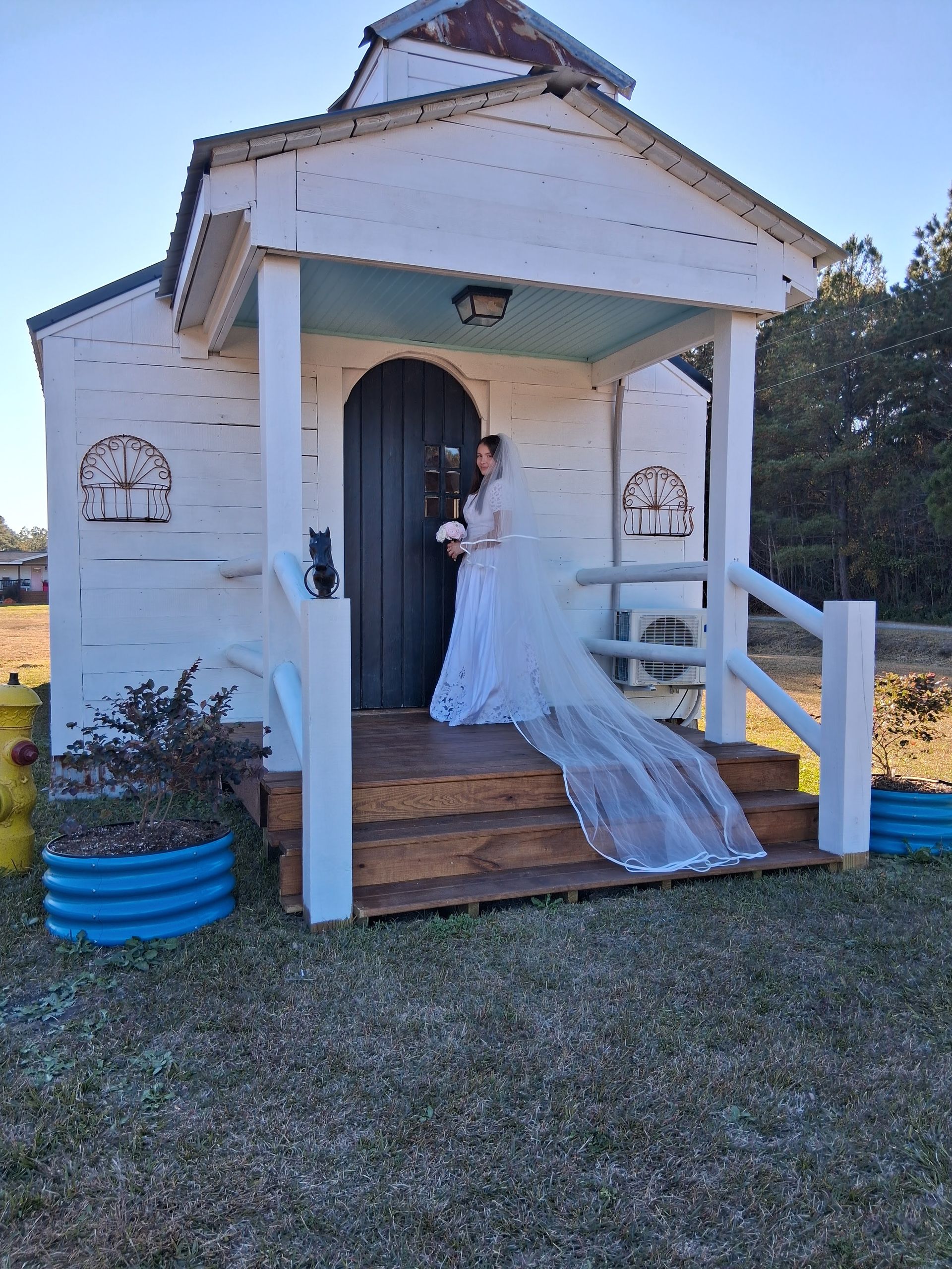 A bride in a long white gown and veil stands at the entrance of a small white wooden chapel under a clear blue sky.
