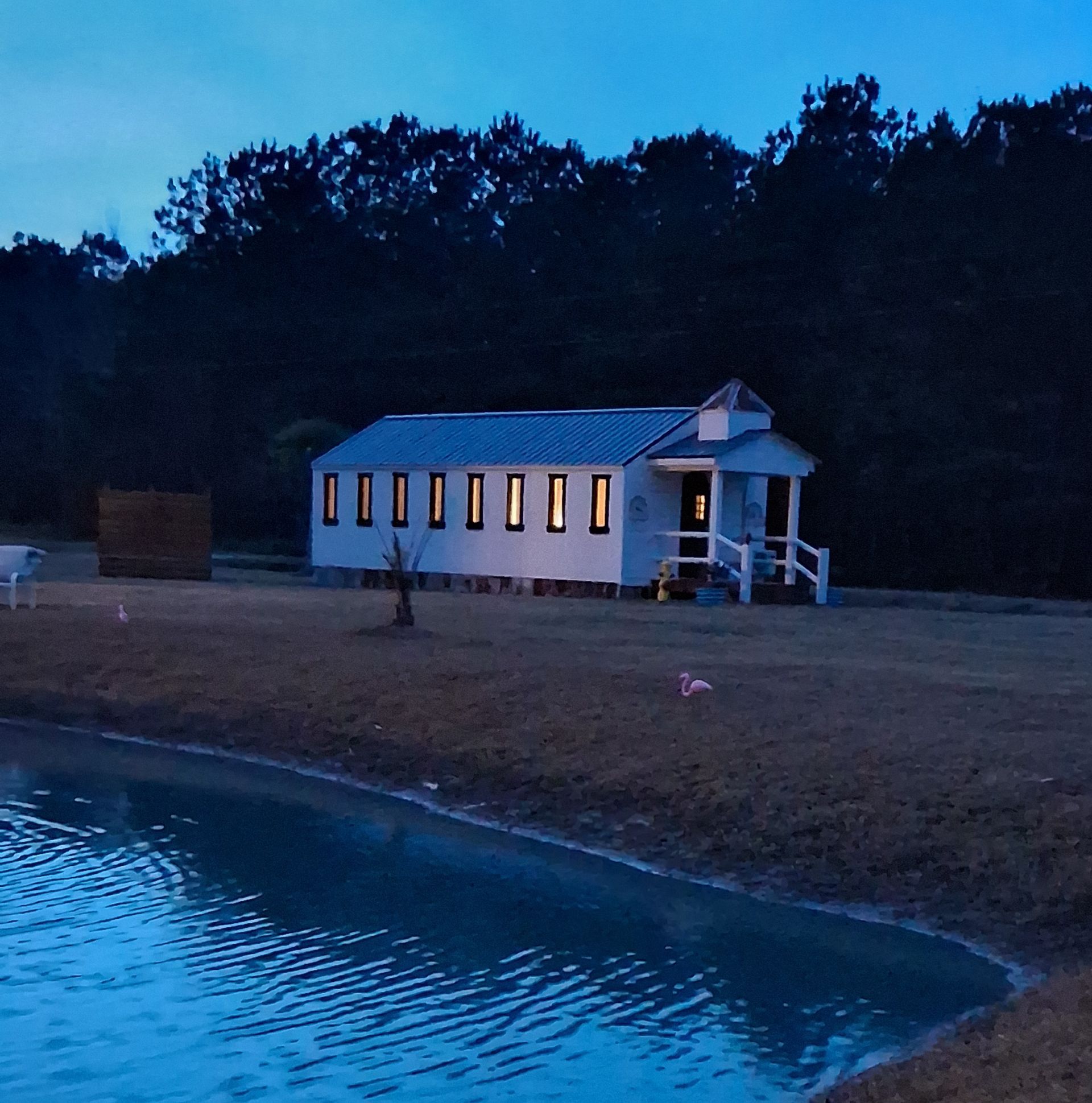 A small white chapel with glowing windows sits near a pond at twilight in front of a line of dark trees. A small white chapel with glowing windows sits near a pond at twilight in front of a line of dark trees.