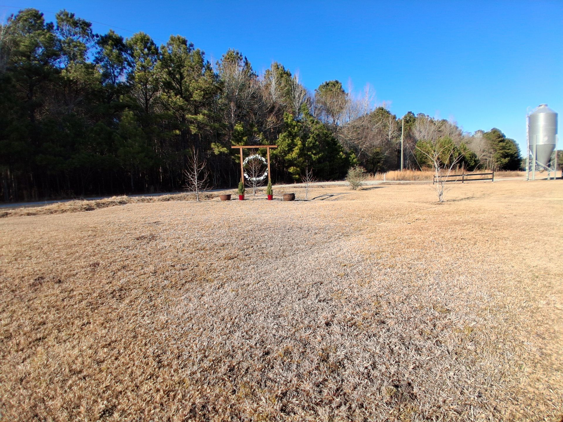 A brown, grassy field leads to a wooden archway decorated with a wreath near a tree line and a metal silo under blue sky.