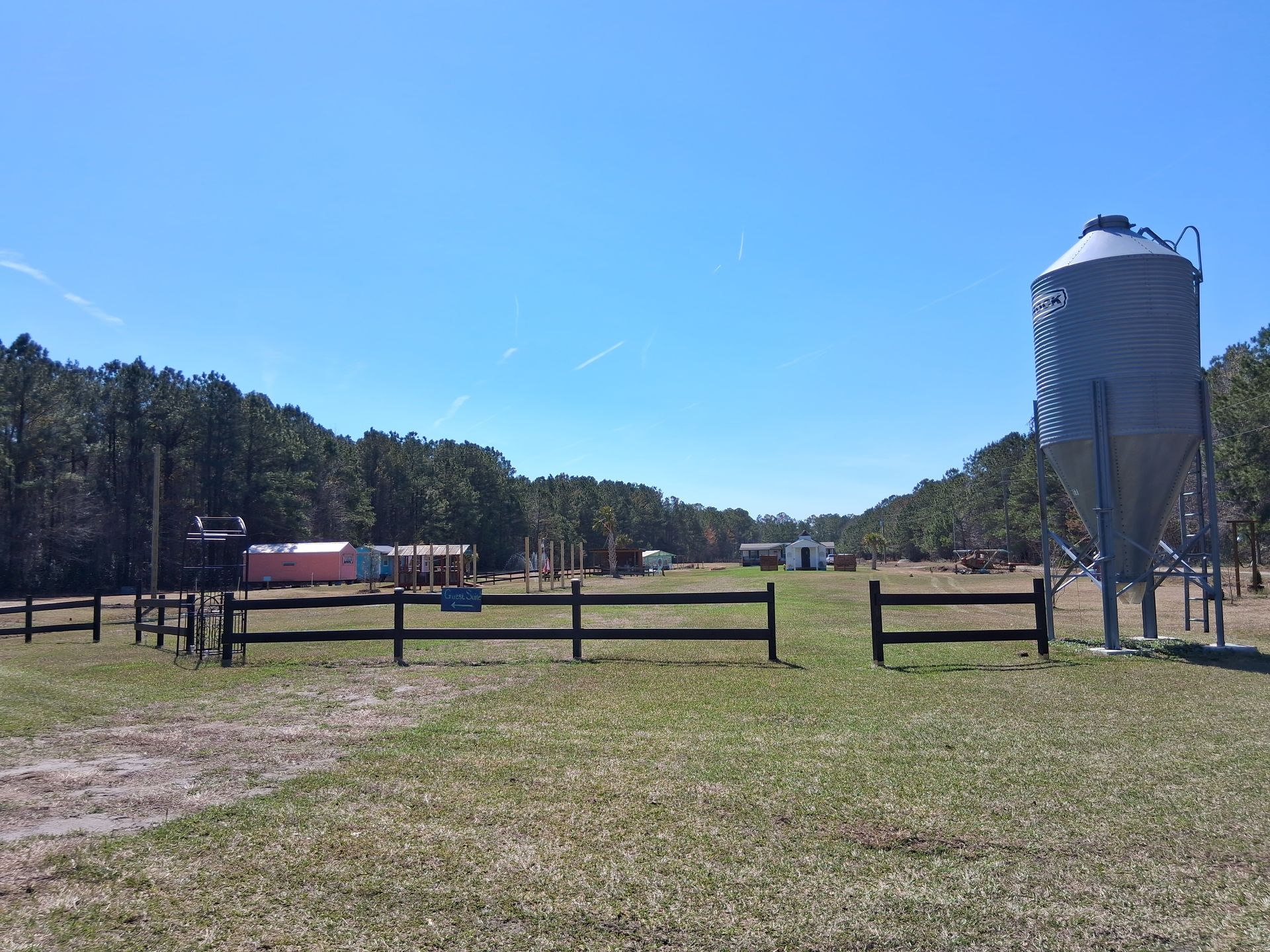 A fenced rural field with a large metal grain silo on the right, trees in the background, and small outbuildings. A fenced rural field with a large metal grain silo on the right, trees in the background, and small outbuildings.