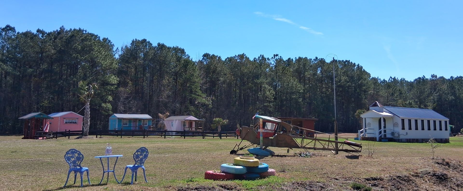 A cluster of small, colorful cottages and a larger white building sit in a grassy field before a treeline. A cluster of small, colorful cottages and a larger white building sit in a grassy field before a treeline.