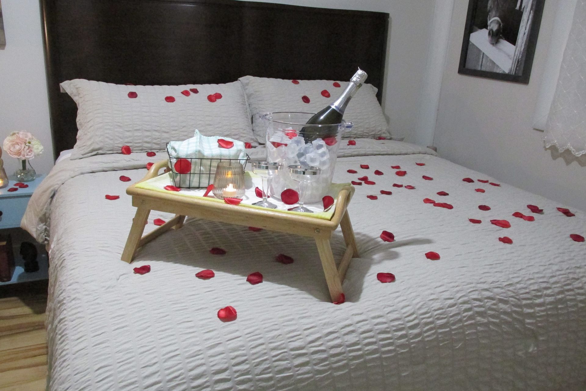 A bed covered in red rose petals, featuring a breakfast tray with champagne and white linens in a romantic setting.