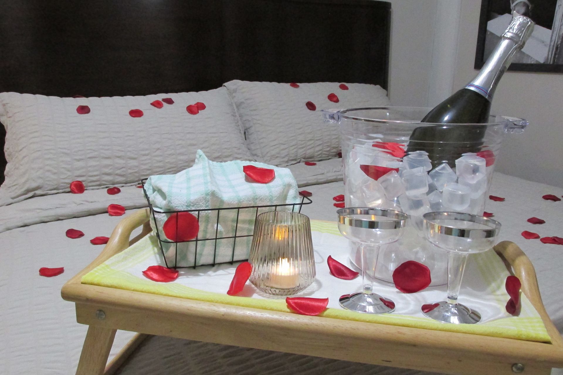 A wooden breakfast tray on a bed features a bottle of champagne in an ice bucket, two glasses, a candle, and rose petals.