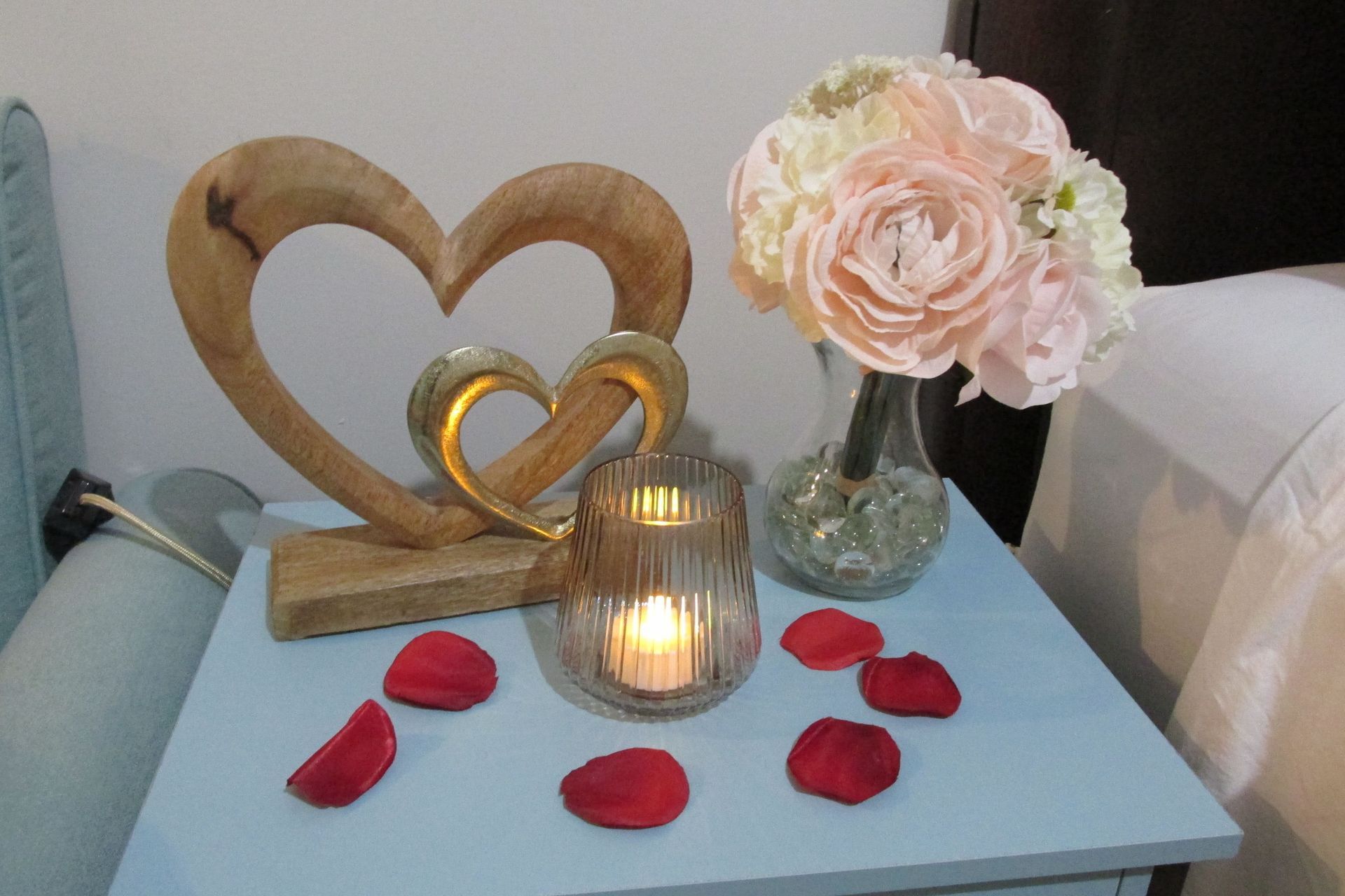 A light blue nightstand with a wooden heart sculpture, a vase of pale pink flowers, a lit candle, and red rose petals.