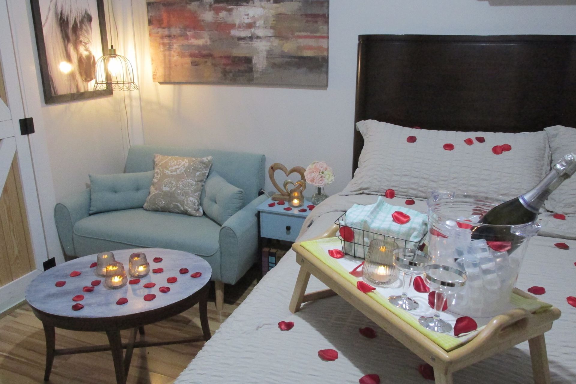 A romantic bedroom setup with rose petals scattered on the bed and round table, candles, and a champagne bottle on a tray.