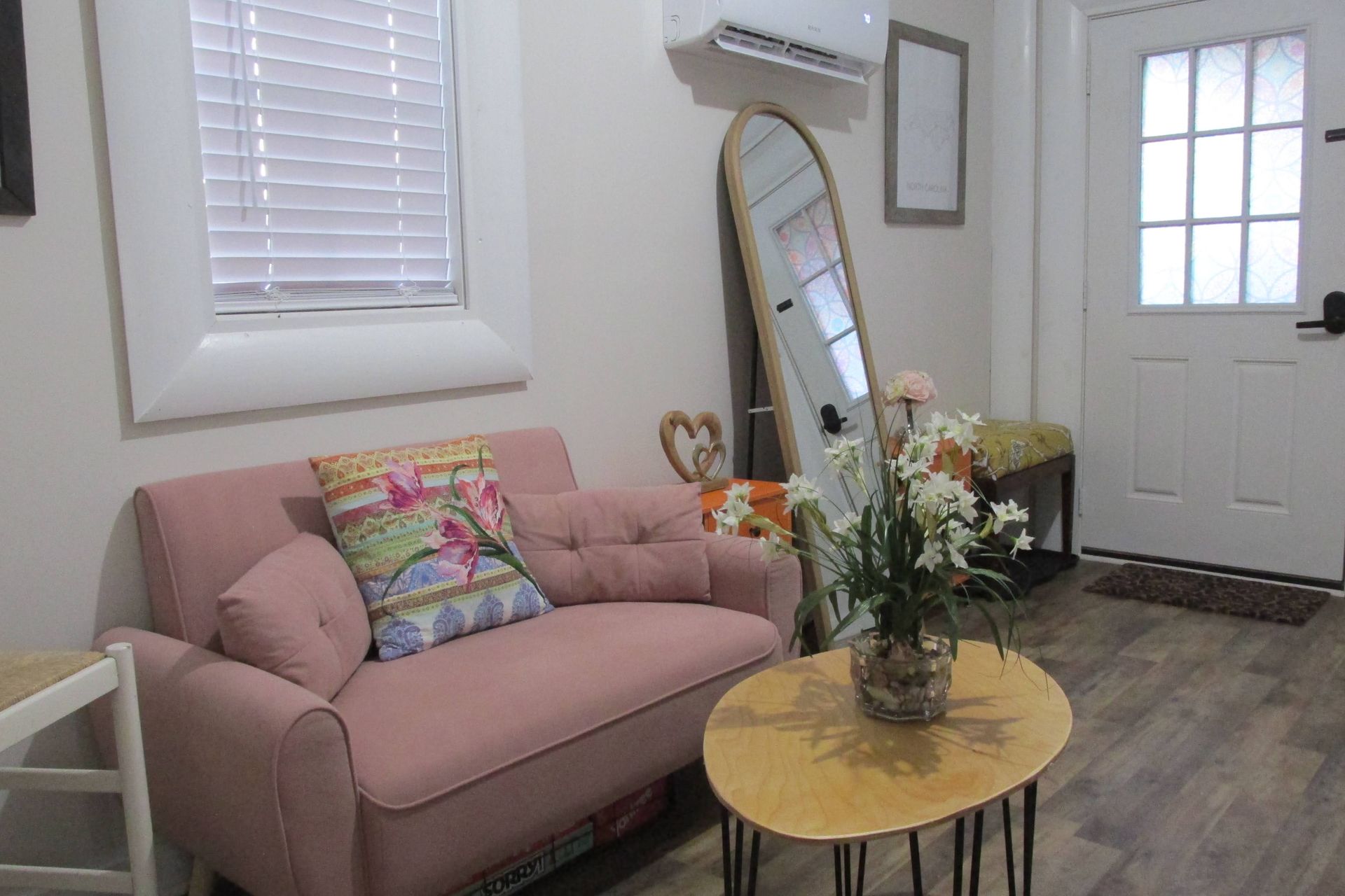 A pink loveseat with a patterned pillow sits next to an oval wooden coffee table, a tall mirror, and a white door.