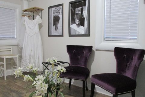A white wedding dress hangs on a rack next to two dark purple tufted chairs, framed horse photos, and white flowers.