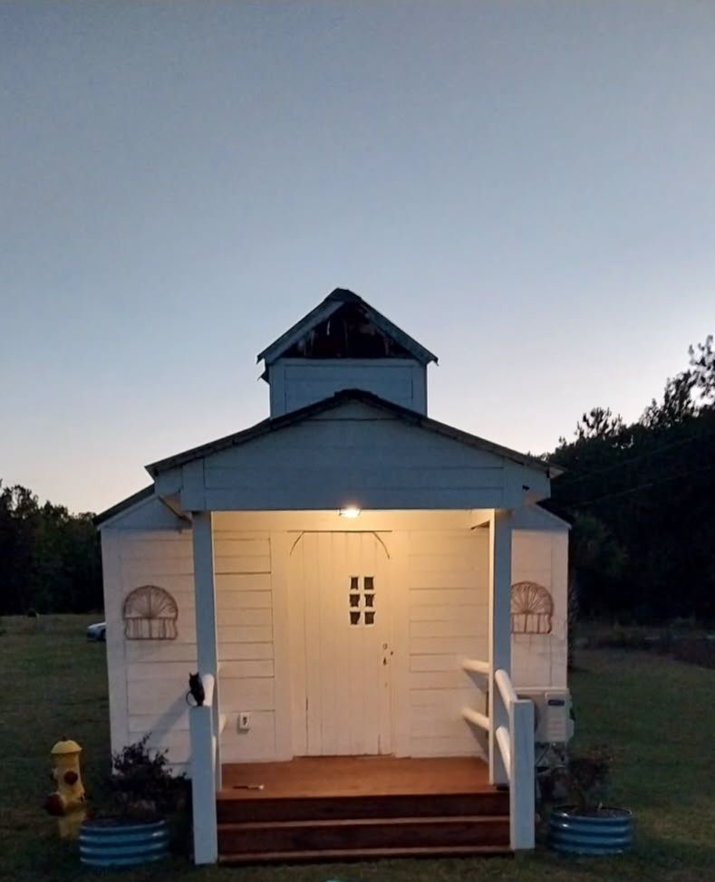 A small white building with a wooden porch and a bell tower stands in a grassy field at twilight.