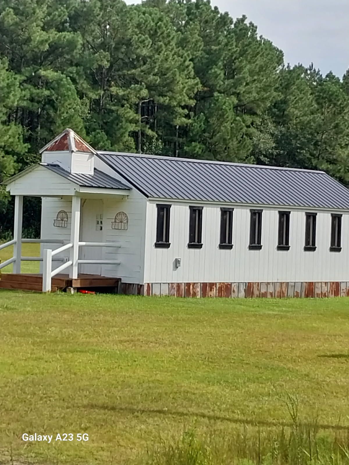 A small, white, rectangular chapel with a dark metal roof and a small steeple sits in a grassy field before a pine forest.
