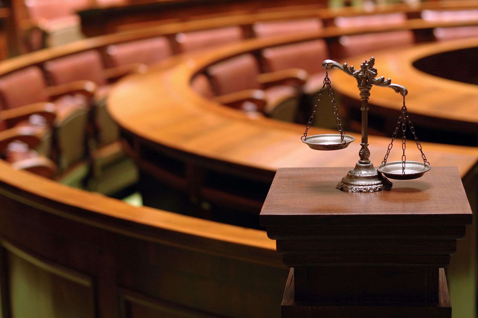 Scales of justice on a wooden pedestal in an empty courtroom with curved wooden seating.