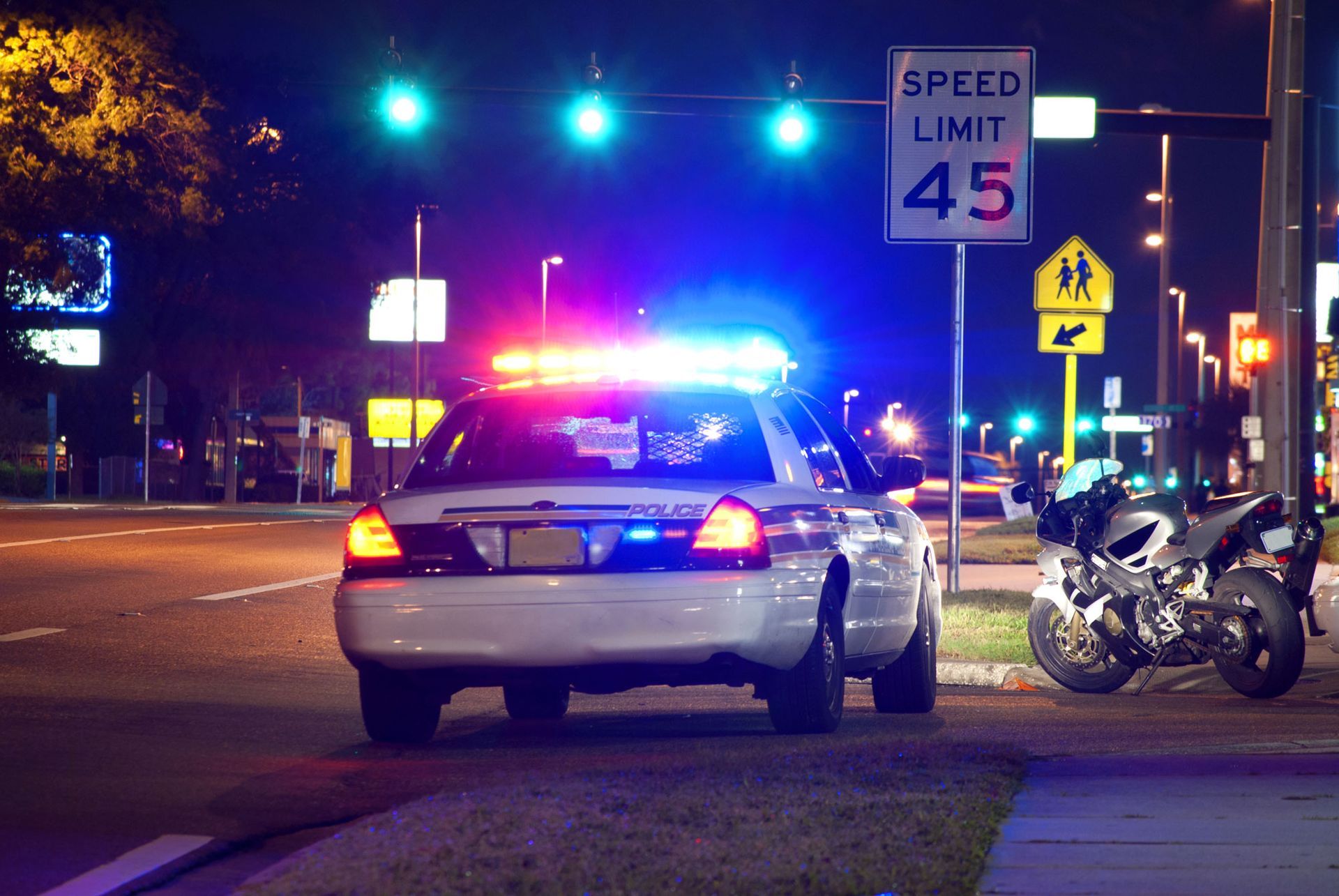 Police car with flashing lights pulled over on a street at night. Motorcycle parked nearby. Speed limit sign.