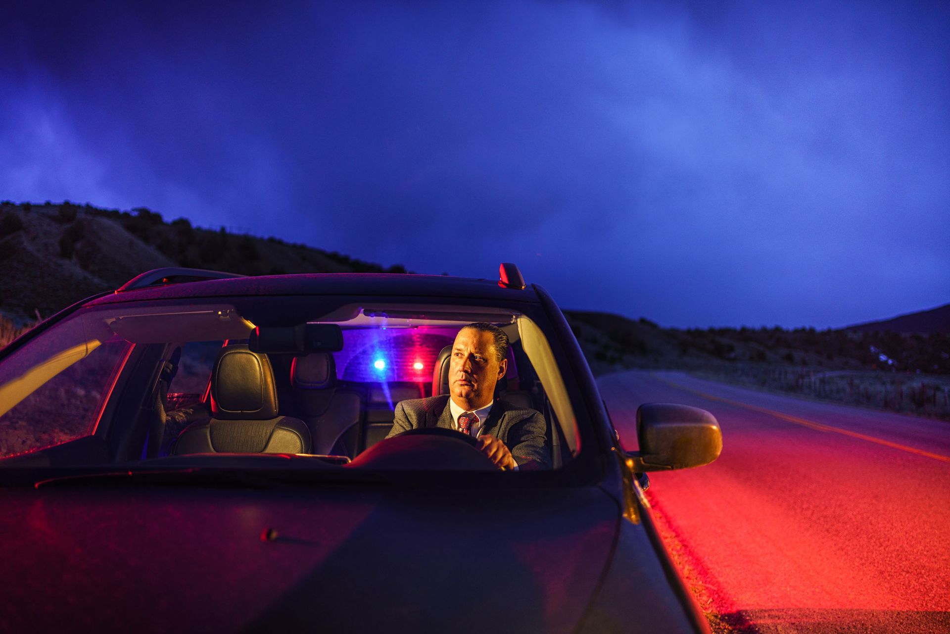 A police officer in a car with flashing red and blue lights on a dark road at night.