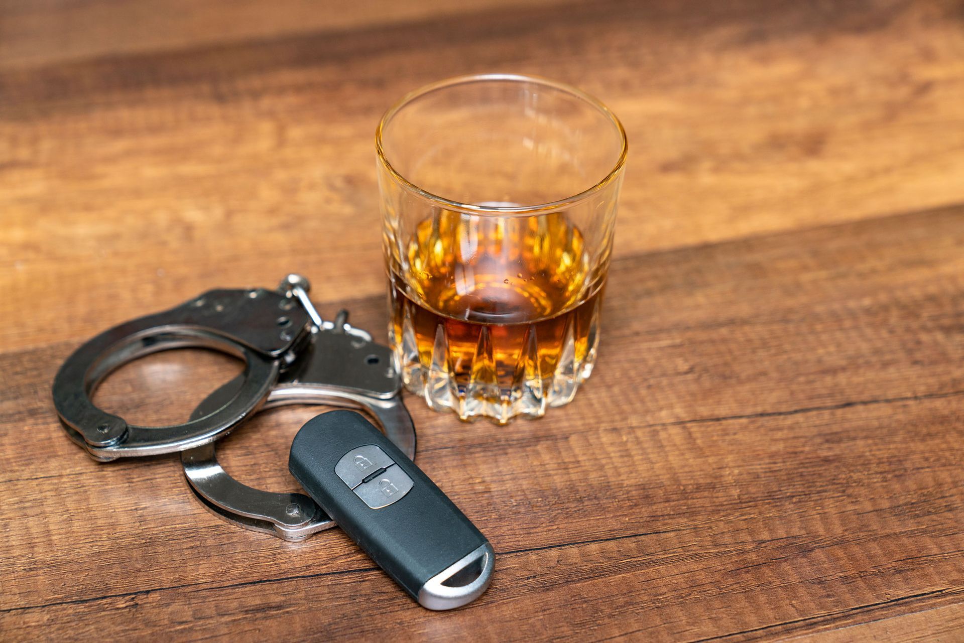 Handcuffs, a glass of whiskey, and a car key on a wooden surface.