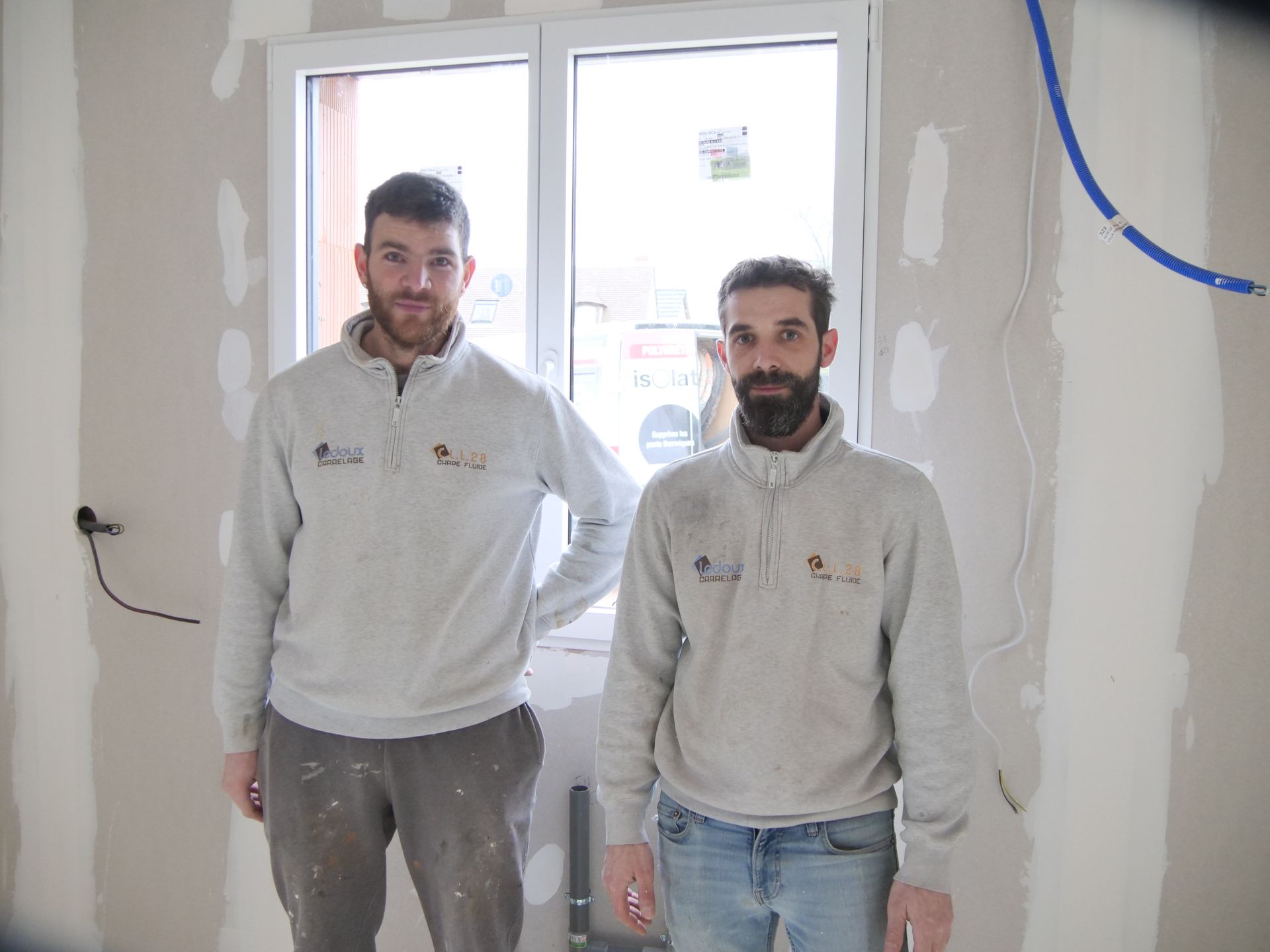 Deux hommes se tiennent l'un à côté de l'autre dans une pièce devant une fenêtre.