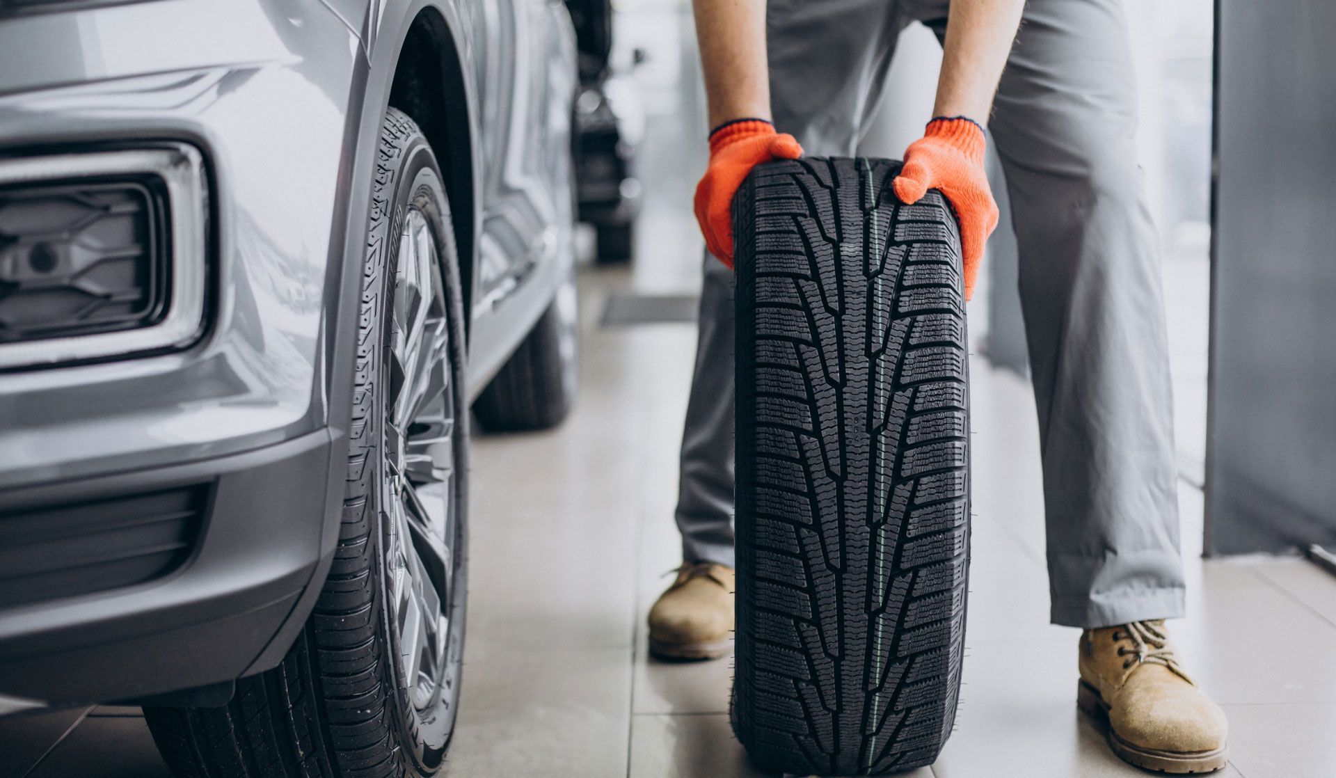 Mechanic holding a tire next to a car in a shop, wearing gloves.