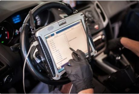 Mechanic using a diagnostic tablet inside a car, wearing gloves, with steering wheel and dashboard visible.