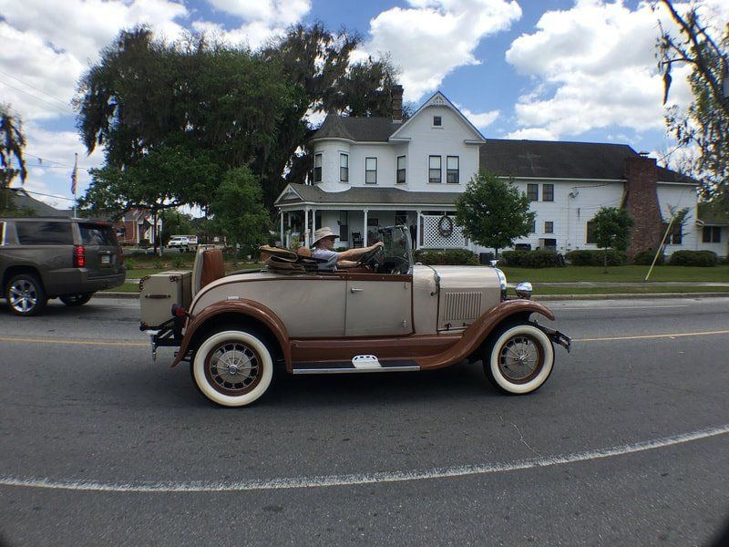 An old car is driving down the road in front of a white house