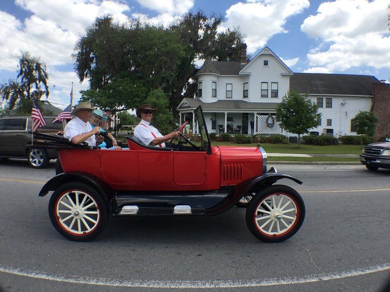 Two men are driving a red car down a street in front of a white house.