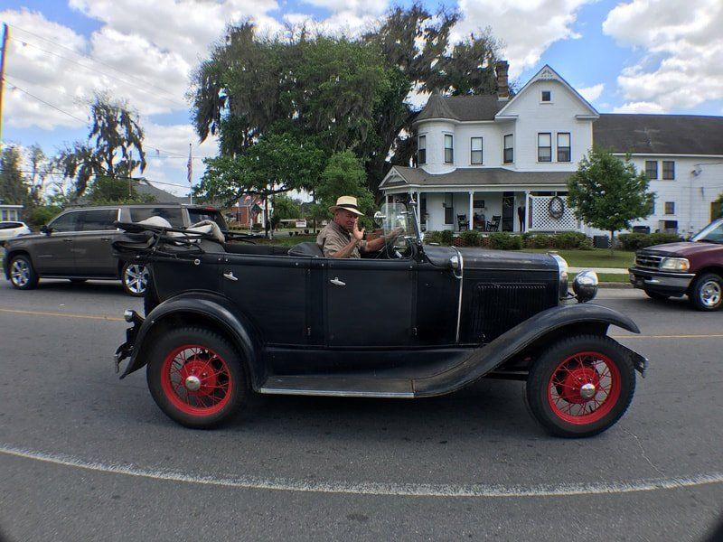 An old black car with red wheels is driving down a street