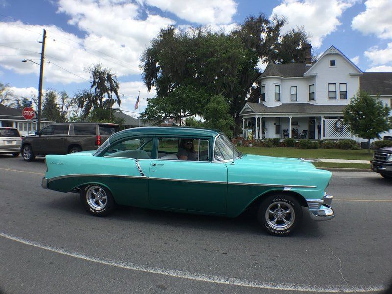 A green and white car is driving down a street in front of a white house.
