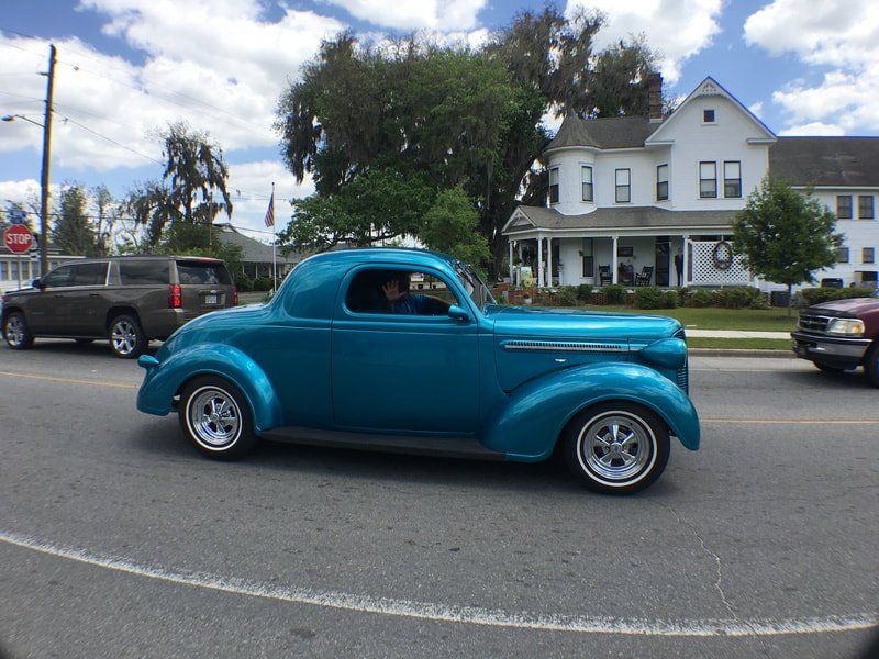 A blue car is driving down a street in front of a white house