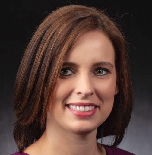 A woman with brown hair is smiling and wearing a purple shirt