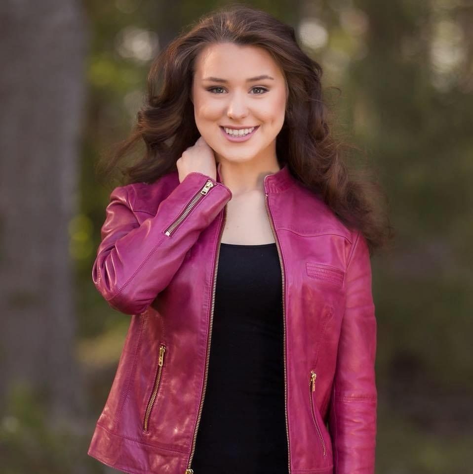 A woman wearing a pink leather jacket and a black dress is smiling.