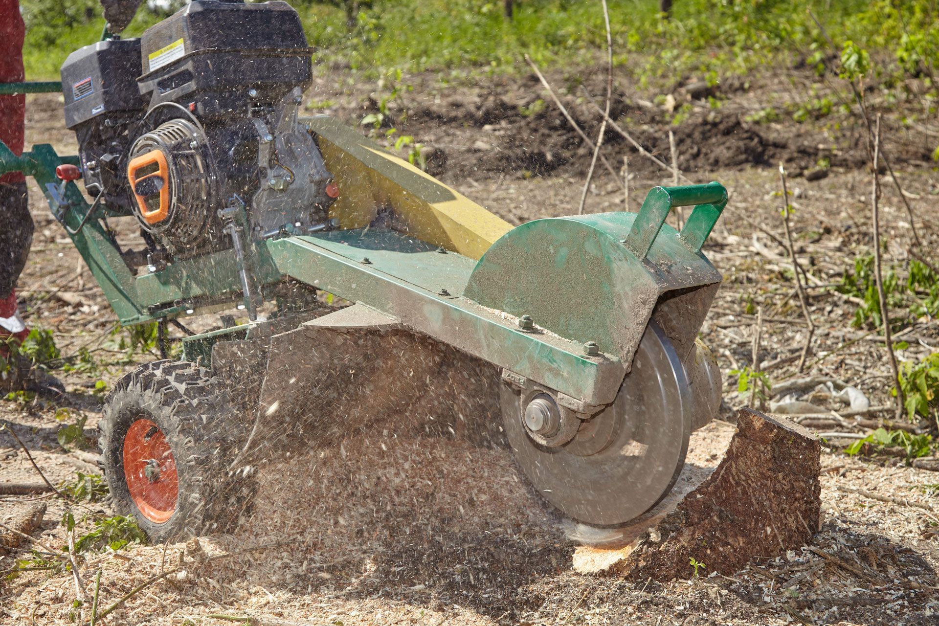 A stump grinder, emitting wood chips, grinds down a tree stump in a field.