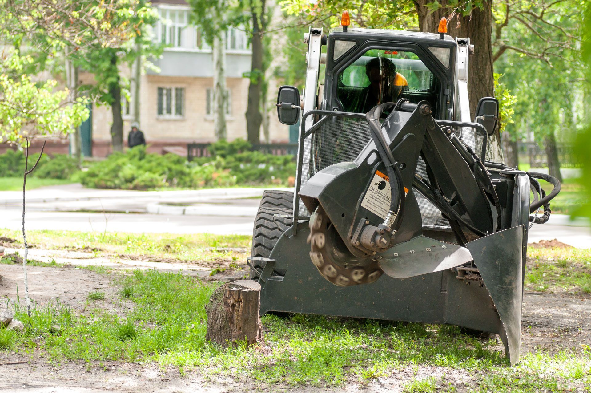 Skid steer stump grinder grinding a tree stump in a grassy area near a sidewalk and building.