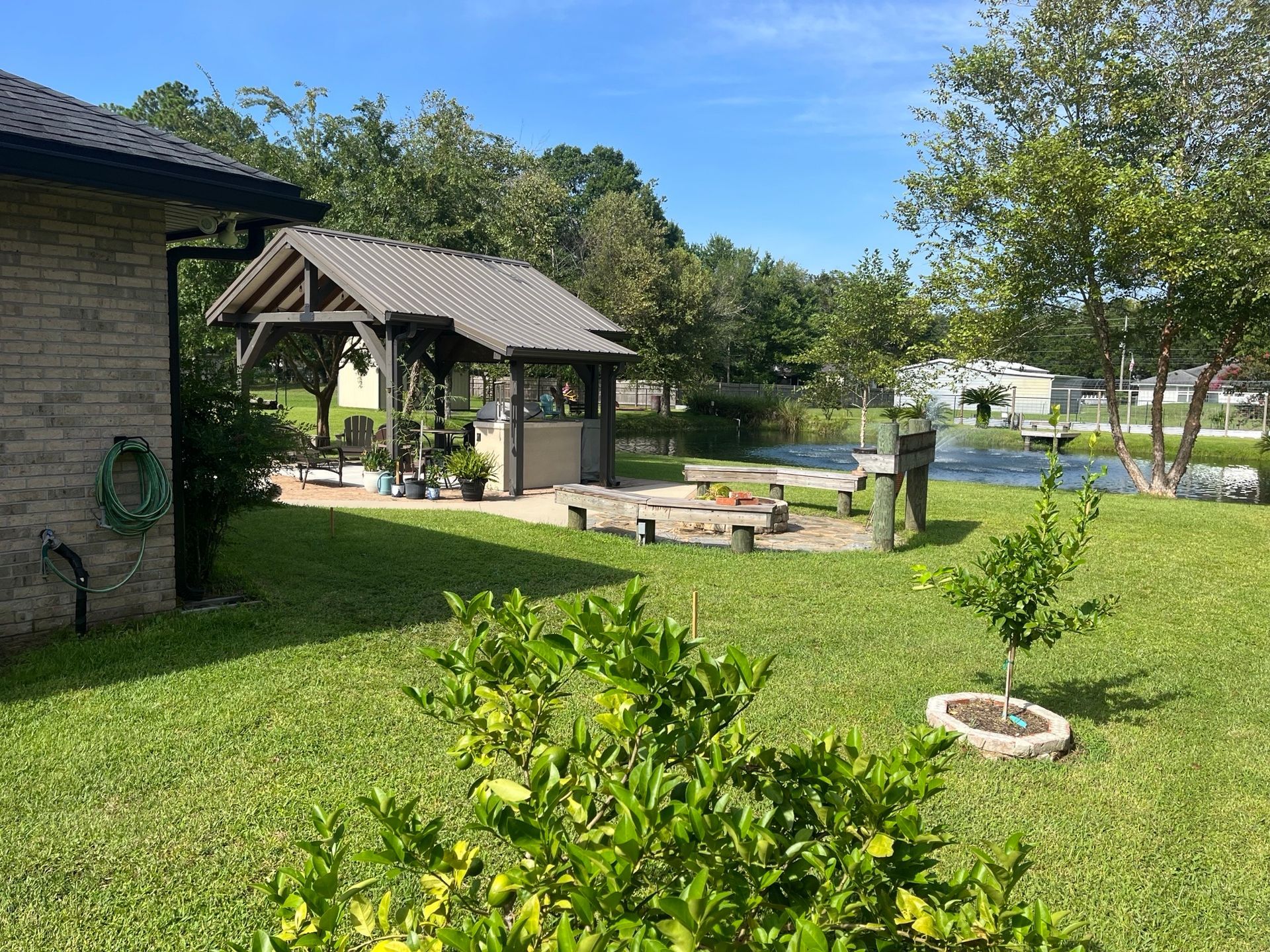 Backyard with gazebo, fire pit, and lawn; lake and trees in the background under blue sky.