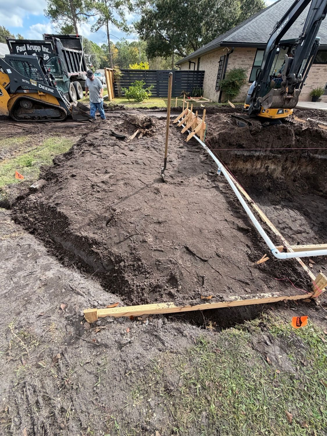 Construction site: Excavation in progress with heavy machinery, pipes, and a worker near a house.