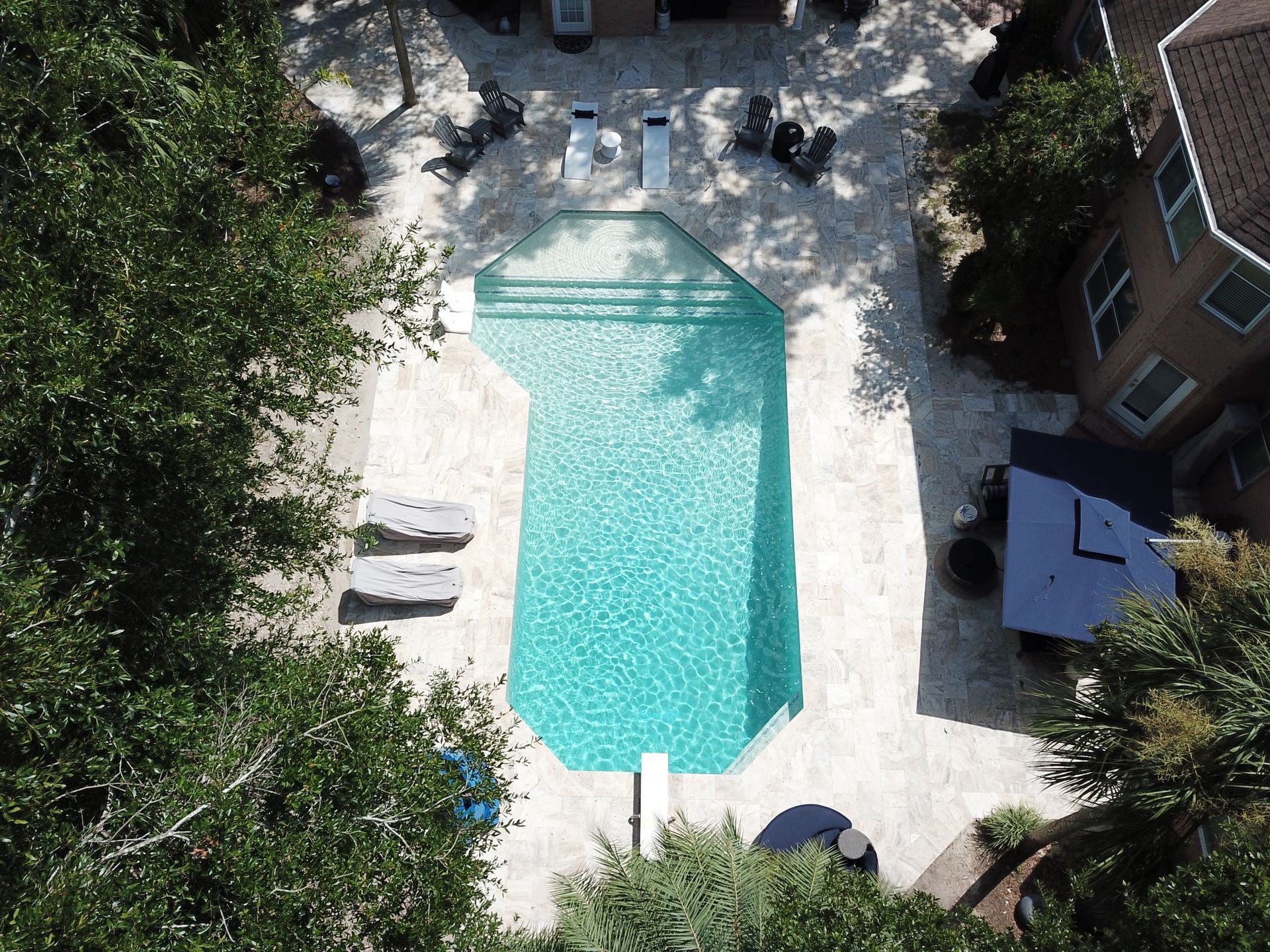 Aerial view of a swimming pool with lounge chairs on a stone patio surrounded by trees and a building.