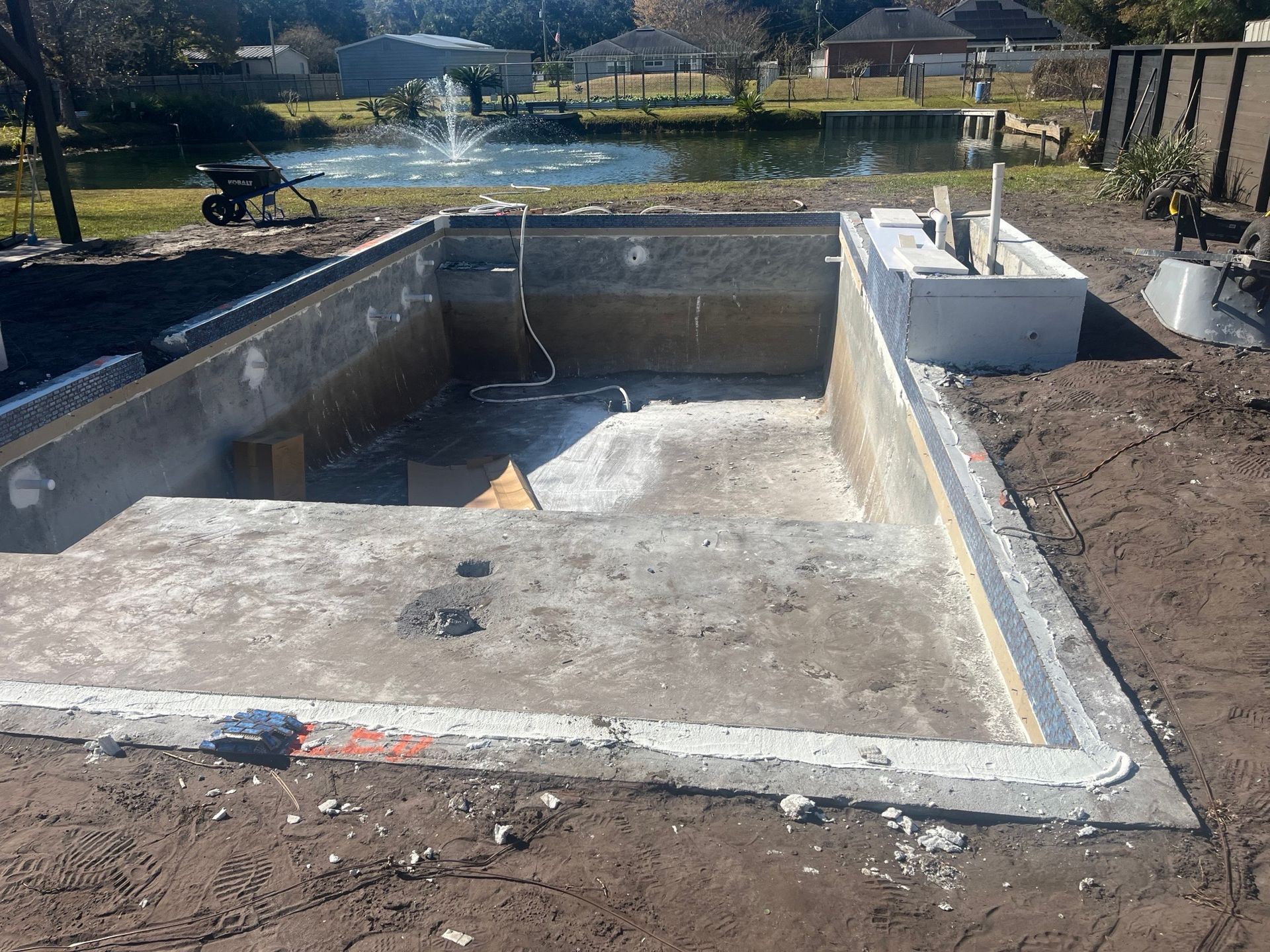 Concrete pool under construction, featuring steps and surrounding ground. In the background, there's a pond with a fountain.