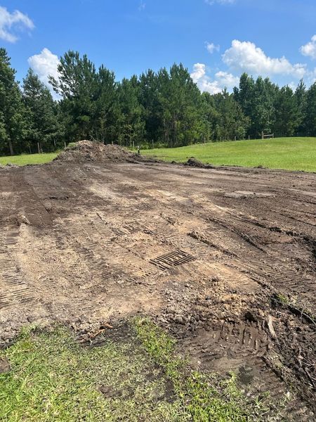 Cleared dirt field with a pile of dirt, green grass, and trees in the background under a blue sky.