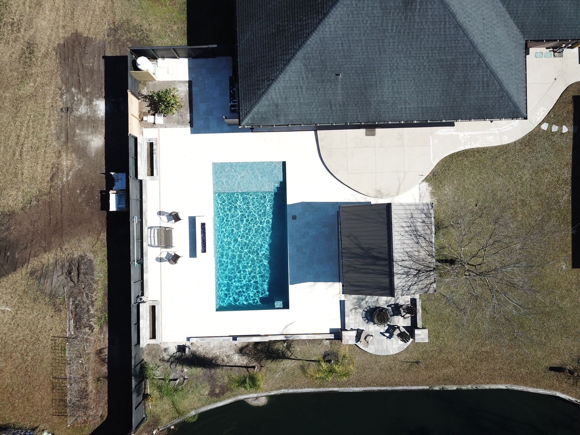 Overhead view of a house with a pool, patio, and dark roof. The pool has blue water, surrounded by light-colored concrete.