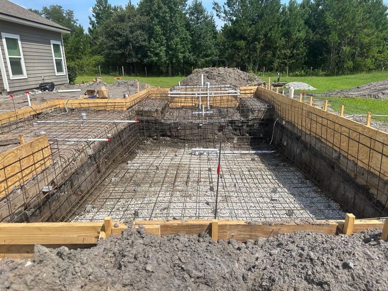 Dirt field with tire tracks and pile of dirt in front of a green grassy area and trees under a blue sky.