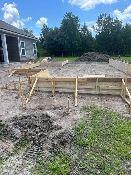 Wooden forms outlining the foundation for a construction project on a dirt lot. A partially built house is in the background.