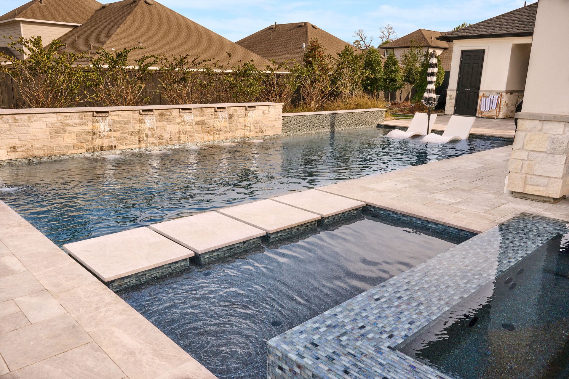Swimming pool with a stone bridge and built-in hot tub, featuring blue tile and stone work, in a backyard setting.