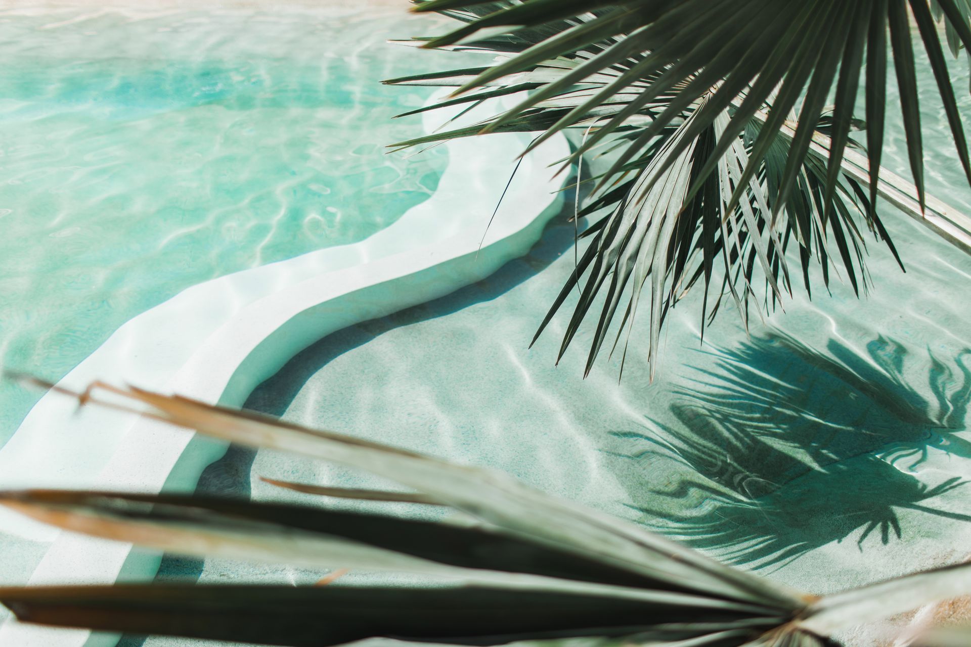 Pool with clear, turquoise water. Palm fronds cast shadows. White pool steps.