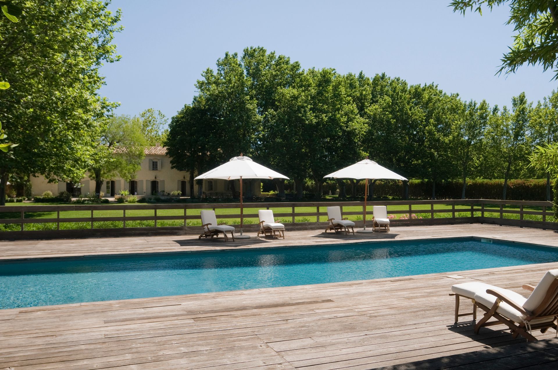 Poolside scene with lounge chairs, umbrellas, and a house in the background. Green trees surround.