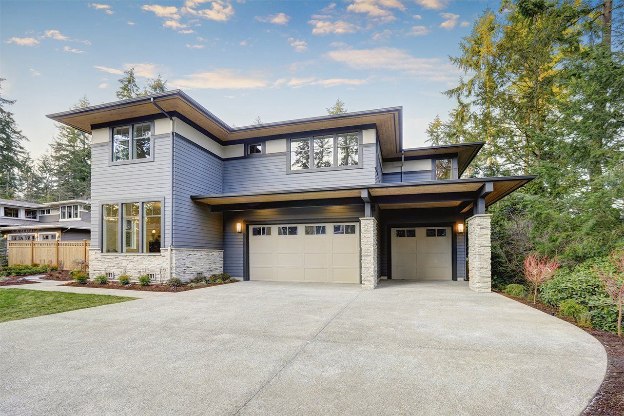 Modern two-story house with blue siding, tan garage doors, and a concrete driveway under a cloudy sky. Modern two-story house with blue siding, tan garage doors, and a concrete driveway under a cloudy sky.