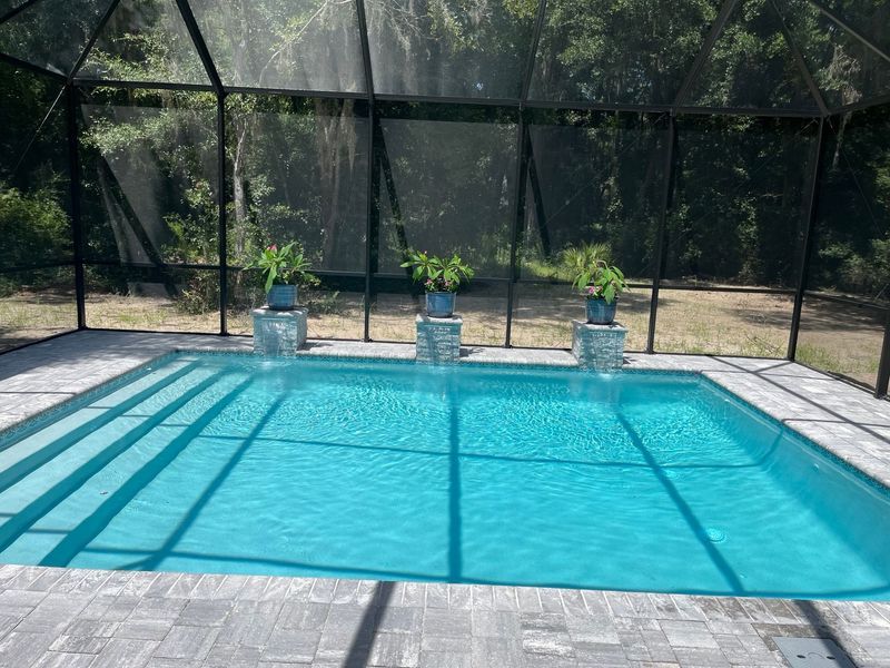 Pool with turquoise water, surrounded by gray pavers and a screened enclosure, with potted plants on pedestals.