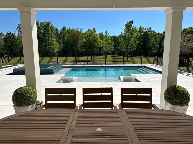 Modern pool with a spa, white sun deck, and a black fence. Trees and blue sky in the background.