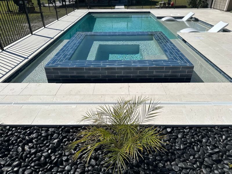 Swimming pool with raised spa, surrounded by decking, black pebbles, and small palm.
