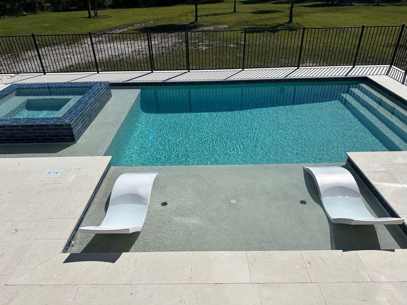 Pool with hot tub, lounge chairs, and steps. Blue water and light-colored decking. Black fence in background.