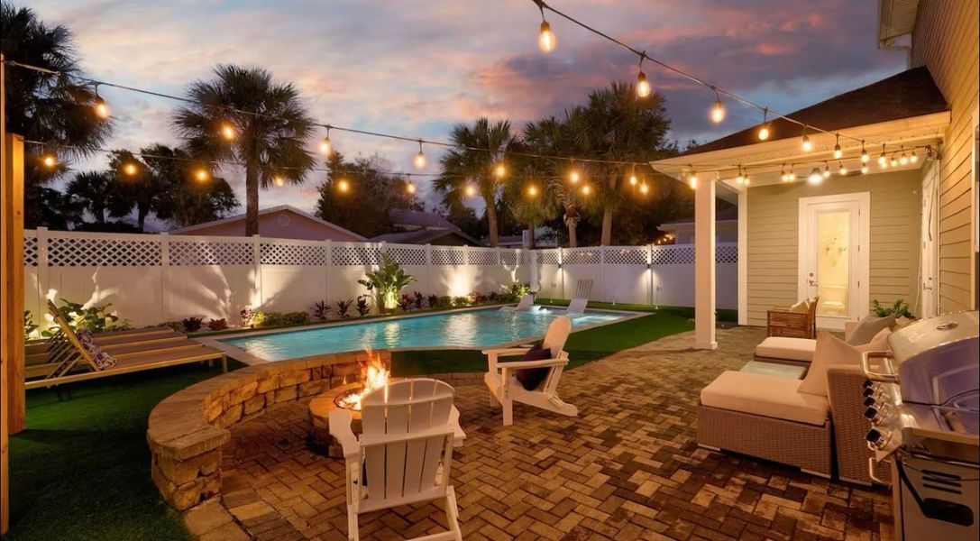 Backyard patio with pool, fire pit, and string lights at dusk.