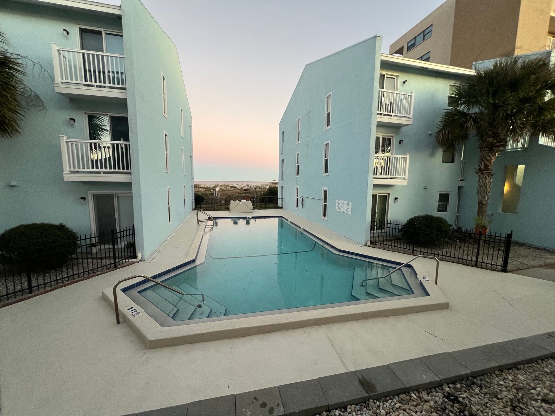 Pool between two turquoise buildings, view of beach beyond.