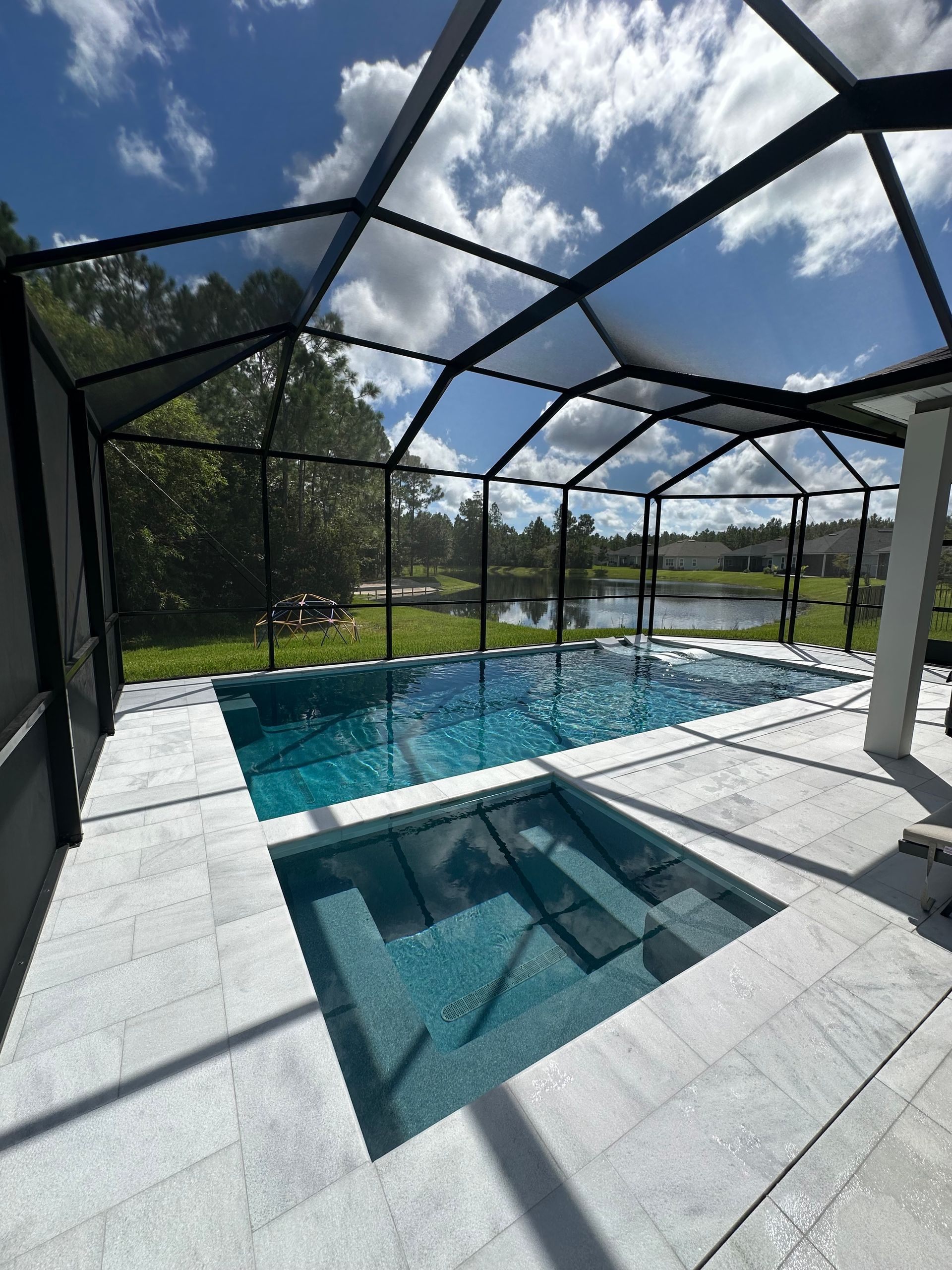 Swimming pool and spa enclosed by screen under a blue sky, surrounded by white stone patio.
