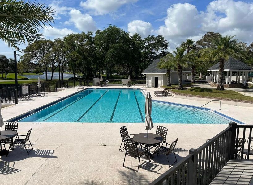 Swimming pool on a sunny day with tables and chairs in the foreground, trees and a building in the background.